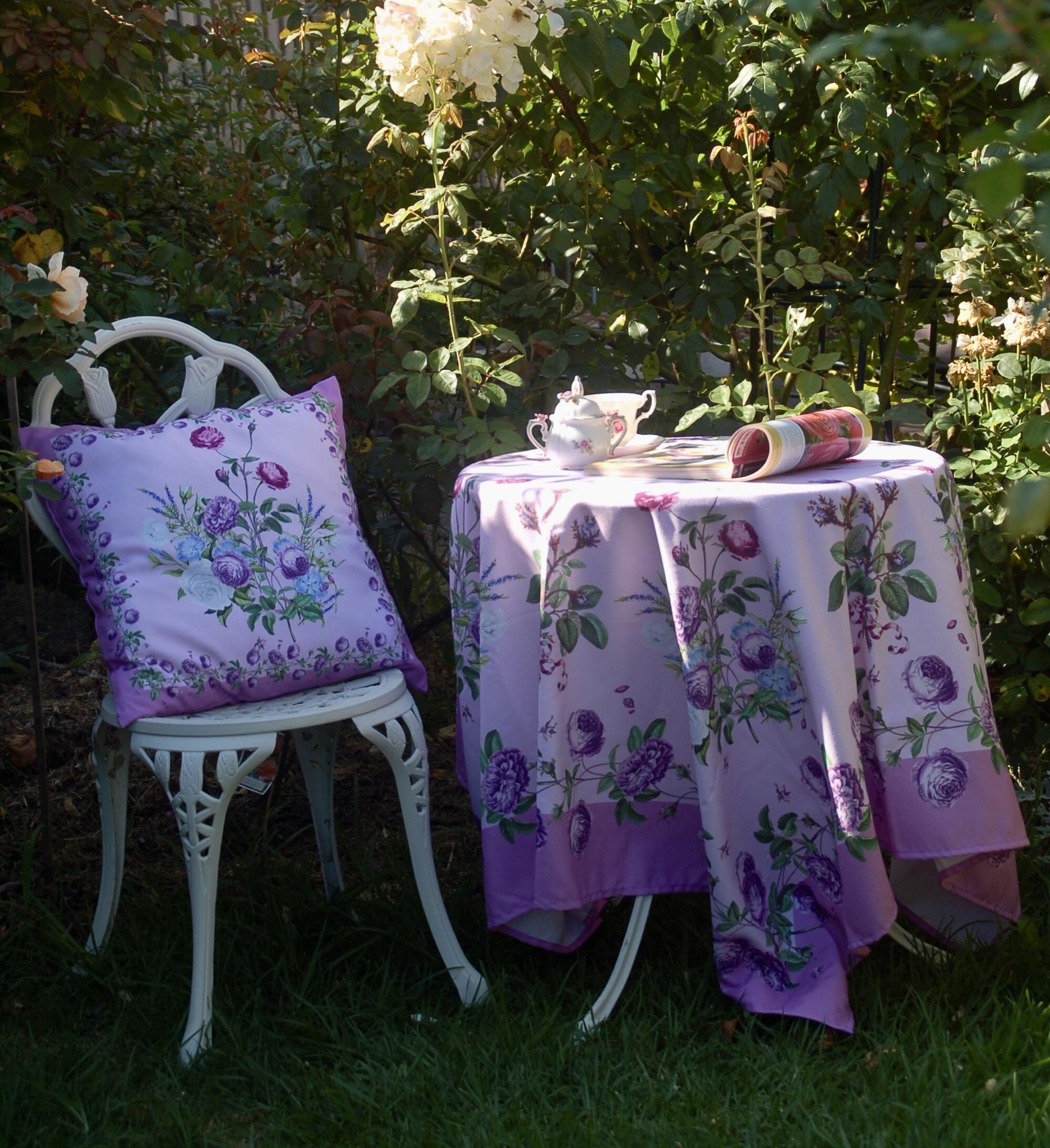 Gối tựa hoa hồng mầu tím phối với khăn trải bàn hoa tím cùng bộ ngoài vườn với tách trà và cuốn sách 